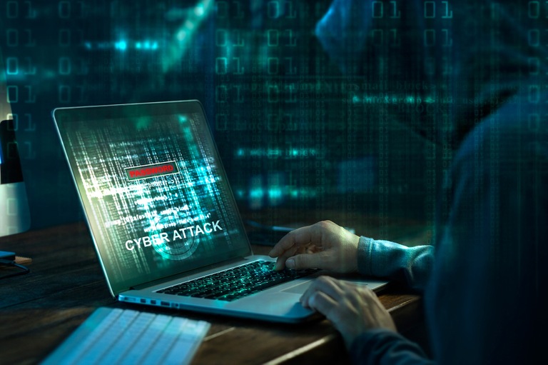 Stock image depicting a hooded computer hacker using a laptop to conduct a cyber attack with code on the screen and in foreground and background of image.